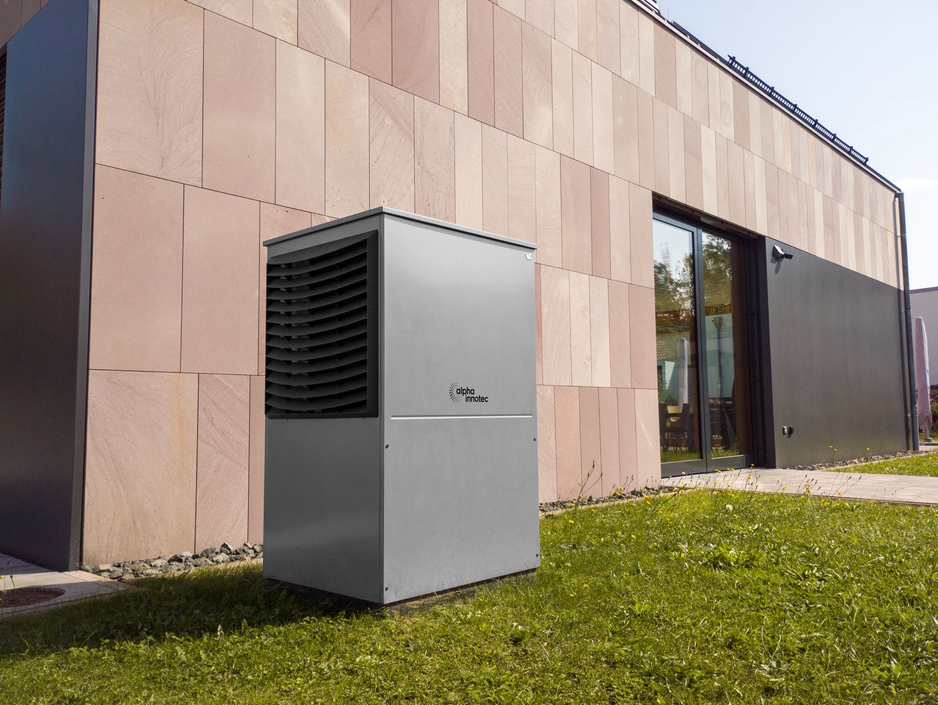 Een grijze buitenunit van een warmtepomp op het gras naast een gevel met roze natuursteen
